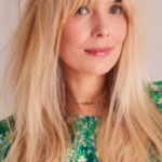 A person with long blonde hair and bangs is wearing a green patterned top and looking at the camera.